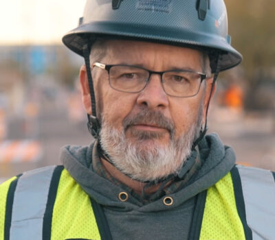 Employee-owner Aric Smith wearing a black hard hat, black-rimmed glasses, a grey hooded sweatshirt, and a high visibility vest.