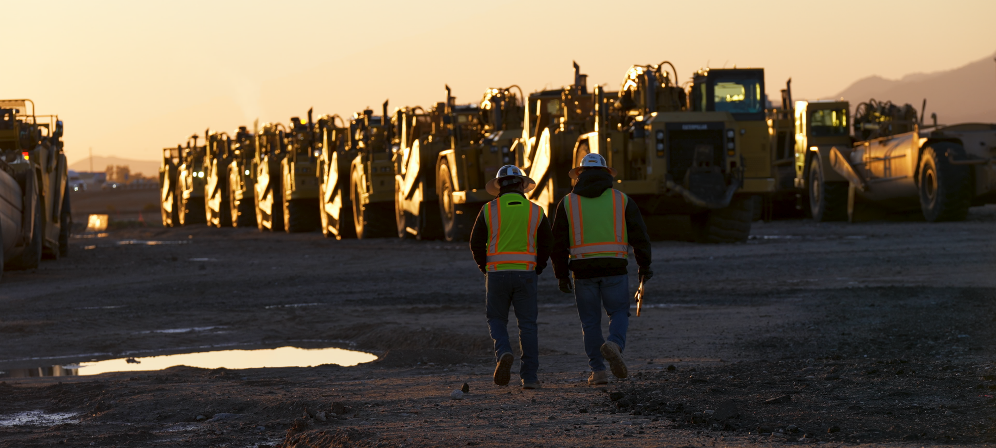Two construction workers wearing hard hats walk side-by-side toward a row of equipment as the sun sets.