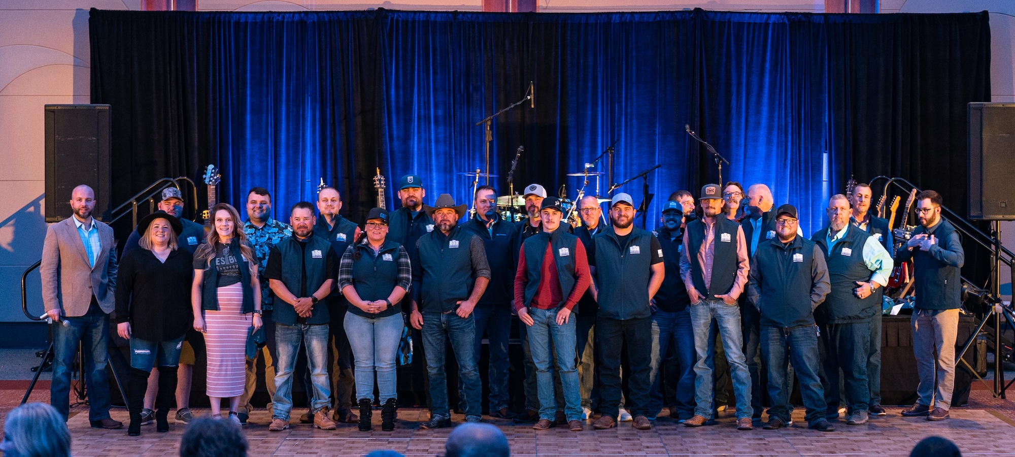 A large group of employees stand in front of a stage, all wearing matching vests to celebrate their fully vested status in the company ESOP program.