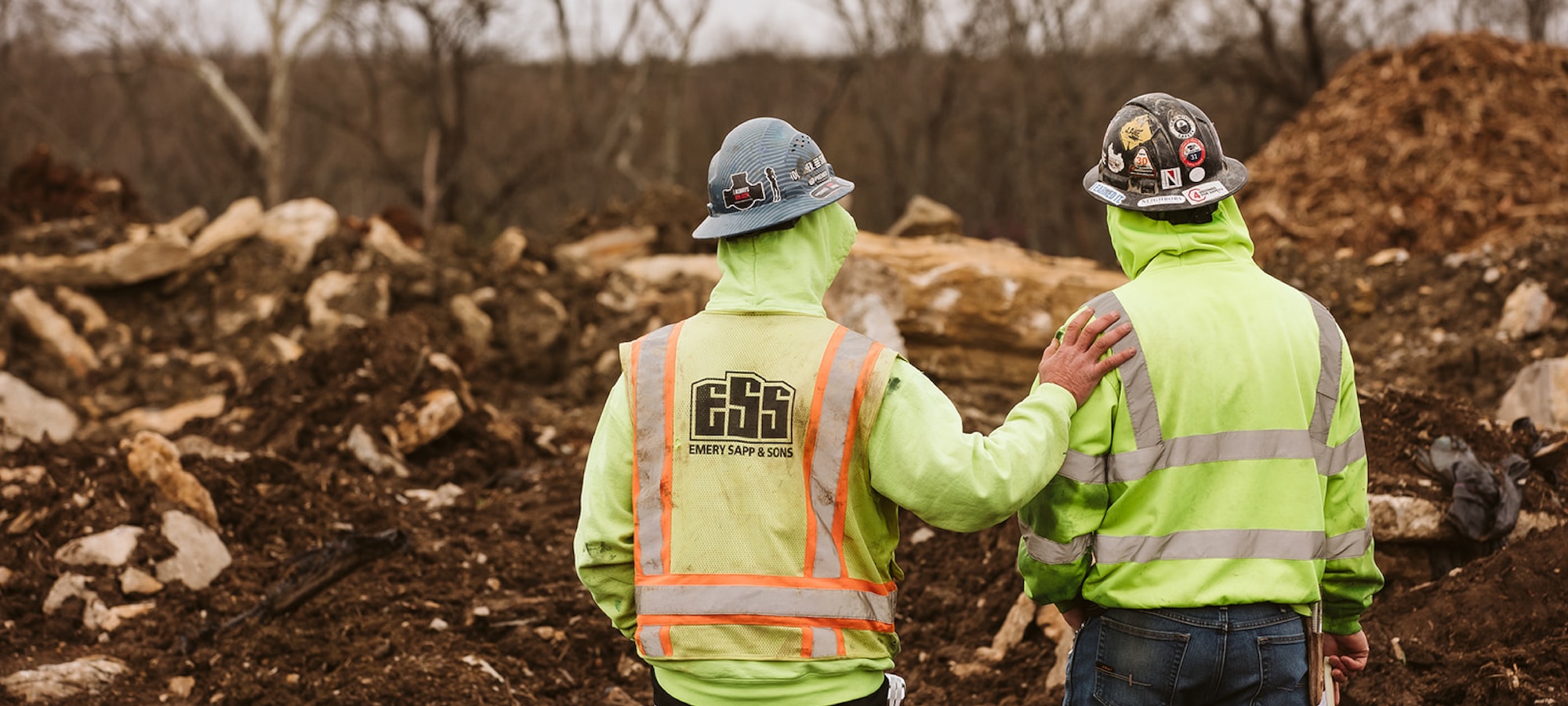 Two construction workers stand with their backs to the camera, looking out at a job site. One man rests his hand on the other man's shoulder.