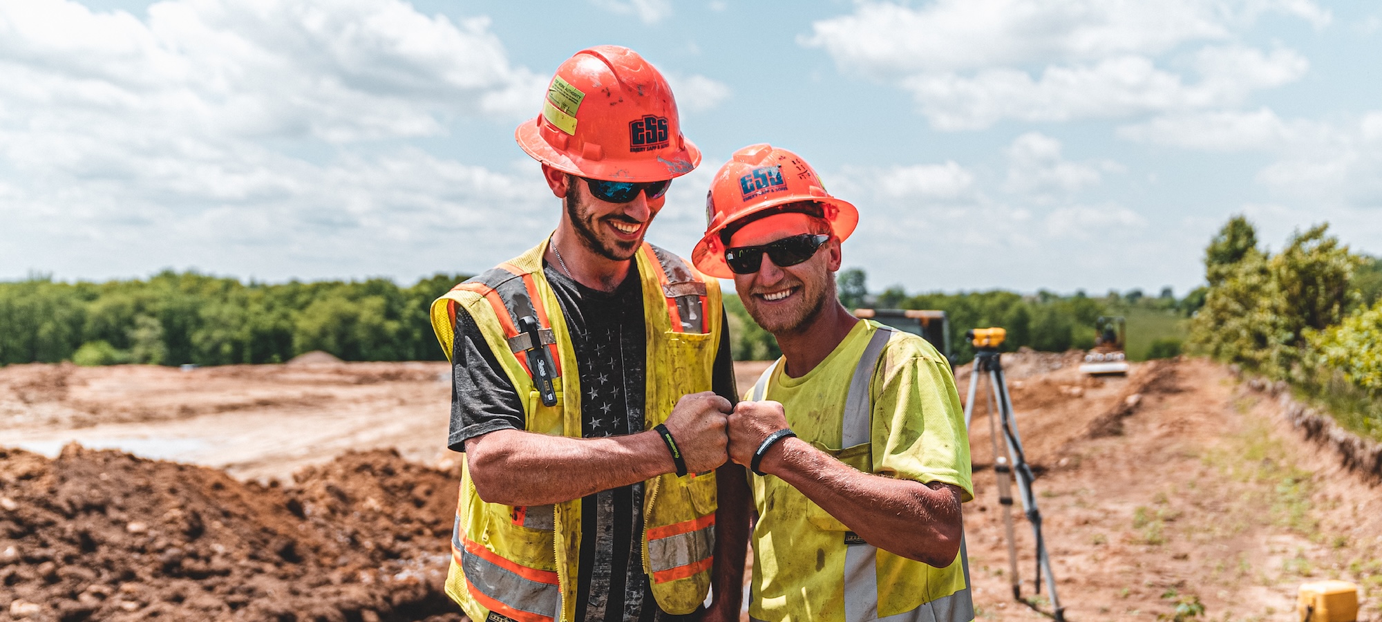 Two male construction workers smile and fist bump in front of a job site. Both are wearing orange hard hats, sunglasses, and high visibility clothing.